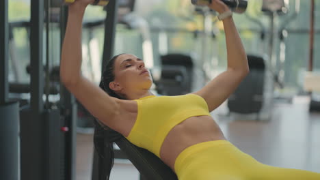 A-young-brunette-woman-in-a-yellow-suit-is-pushing-a-dumbbell-while-lying-on-the-bench.-Dumbbell-bench-press.-Exercise-for-chest-muscles